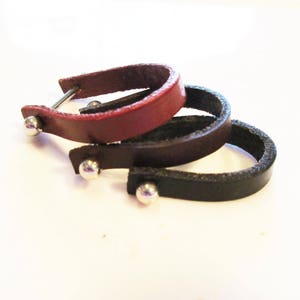 May include: Three leather bracelets with silver metal accents. The bracelets are stacked on top of each other, with the black bracelet on the bottom, the brown bracelet in the middle, and the red bracelet on top.