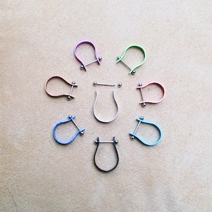 Peut inclure: Un ensemble de huit bijoux de barre en forme de fer à cheval de couleurs différentes avec des billes argentées à chaque extrémité. Les bijoux sont disposés en cercle sur une surface beige.