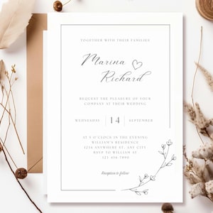 May include: A white wedding invitation with a minimalist design. The text includes the names "Marina & Richard", date, time, and location. Delicate floral illustrations and a simple frame enhance the elegant aesthetic. A brown envelope and dried flowers are in the background.