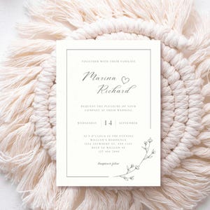 Peut inclure: Une invitation de mariage crème avec les noms "Marina" et "Richard" en écriture élégante. L'invitation comprend la date, l'heure et le lieu du mariage, avec des accents floraux. L'invitation est posée sur un fond texturé blanc cassé.