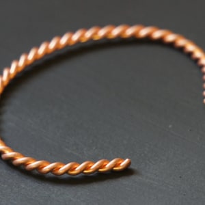 Puede incluir: Un brazalete de alambre retorcido de color cobre. El diseño de brazalete abierto presenta un patrón trenzado continuo. El brazalete está hecho de un material metálico brillante y está colocado sobre un fondo gris oscuro.