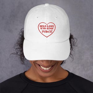 Pode incluir: Boné de beisebol branco com aba curva. O boné apresenta o texto "SELF-LOVE IS MY SUPER POWER" dentro de um coração vermelho. O chapéu é feito de um material macio e é adequado para o uso diário.