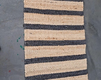 May include: A rectangular jute rug with alternating horizontal stripes of natural beige and dark blue. The rug is made of woven fibers and has a simple, modern design. The rug is suitable for indoor use.