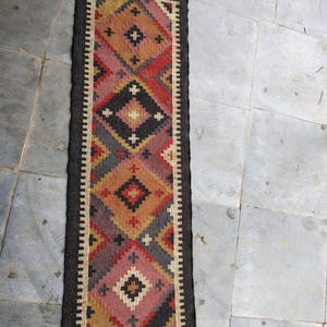 以下が含まれることがあります： 赤、ピンク、黄色、グレーの色合いの幾何学的なひし形模様の長方形の織りランナーラグ。ラグは濃い茶色の縁と両端にフリンジが付いています。ラグの幅は約60cmです。