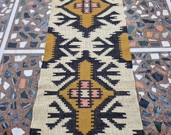 Puede incluir: Una alfombra rectangular con un patrón geométrico. La alfombra presenta un fondo color crema con formas geométricas negras, doradas y rosas. La alfombra mide aproximadamente 152 cm de largo.