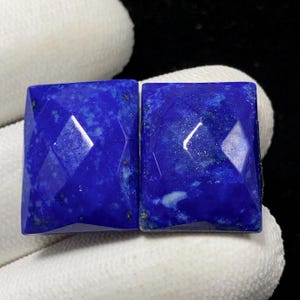 May include: Two square-cut, faceted lapis lazuli gemstones. The gemstones are a deep, rich blue color with subtle variations in tone and small flecks. The facets create a geometric pattern, reflecting light and adding depth to the stones. These gemstones are ideal for jewelry making.