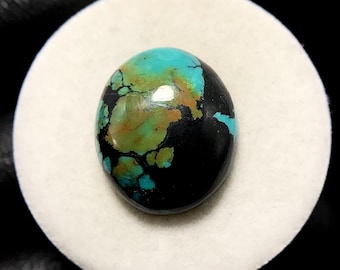 May include: Oval turquoise gemstone with a polished surface. The stone displays a mix of colours, including vibrant turquoise, earthy brown, and deep black, creating a unique pattern. The gemstone is set on a white surface.