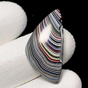 May include: A polished, teardrop-shaped stone with a vibrant, layered pattern of colours. The stone features bands of black, white, red, yellow, and blue, creating a unique, abstract design. The stone is held against a white, textured surface.