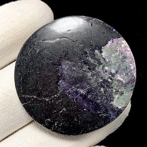 May include: A circular polished gemstone cabochon featuring a deep black base color with prominent inclusions of vibrant purple and mossy green mineral patterns across one side, displaying a smooth, reflective surface texture throughout.