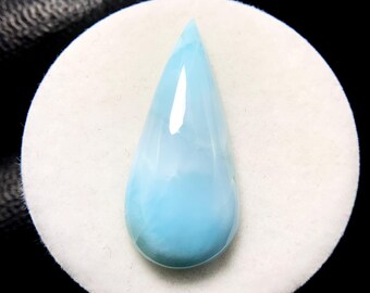 May include: A teardrop-shaped, polished gemstone in shades of light blue and white, resting on a white circular surface. The gem has a smooth, glossy finish, and the background is black.