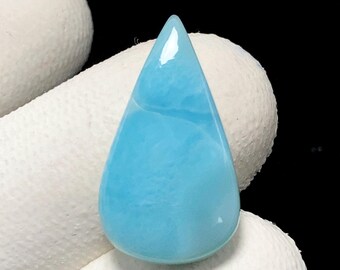 May include: A teardrop-shaped, polished gemstone in a light blue hue, reminiscent of ocean water. The stone has a smooth, glossy surface and a slightly translucent appearance. The gemstone is held against a white background.