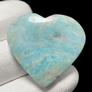 May include: A heart-shaped gemstone in shades of light blue, featuring natural veining. The polished stone has a smooth texture. The heart is held by a gloved hand, set against a black background.