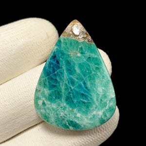 May include: A teardrop-shaped gemstone featuring a vibrant mix of teal and turquoise hues with intricate white veining throughout. The top section displays a small, translucent, brownish-tan crystalline inclusion, while the rest of the stone exhibits a polished, smooth surface with a natural, marbled texture.