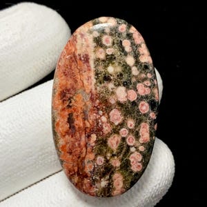 May include: Oval-shaped gemstone with a unique pattern. The stone features a blend of colors, including reddish-brown, olive green, and pink spots. The polished surface highlights the natural variations and textures of the stone. A beautiful, natural stone for jewelry making.