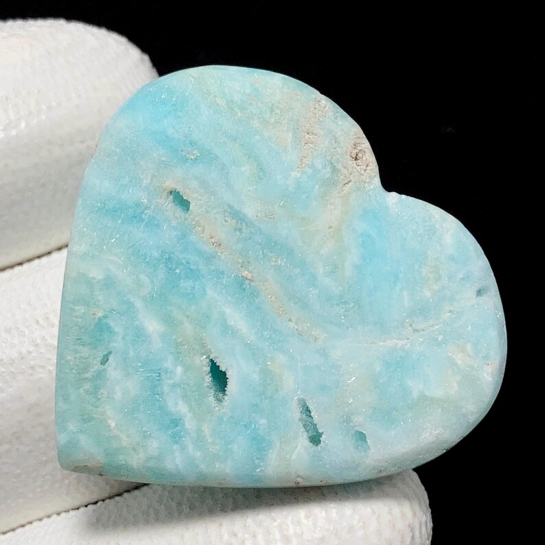 May include: A heart-shaped gemstone in shades of light blue, featuring natural patterns and a polished surface. The stone has small holes and a slightly rough texture. The gemstone is held against a white background.