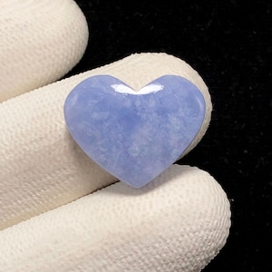 May include: A heart-shaped cabochon made of light blue chalcedony with a smooth, polished surface. The stone features a translucent appearance with subtle, cloudy white inclusions throughout its pale blue color profile.