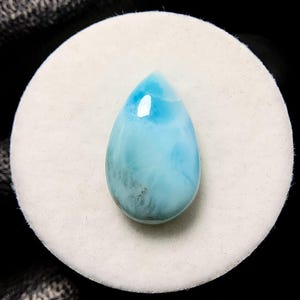 May include: A teardrop-shaped, polished gemstone in shades of light blue and turquoise. The stone has a smooth, glossy surface with subtle variations in color and a few darker inclusions. It is set against a white, circular background.