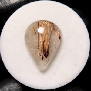 May include: A teardrop-shaped gemstone with a clear, translucent body. The stone features striking inclusions of golden-brown, needle-like rutile crystals, creating a unique, textured appearance. The gemstone is set against a white background.