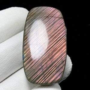 May include: A polished, rectangular gemstone with a rounded top. The stone features a mix of colors, including purple, pink, and gray, with diagonal striations across its surface. The gemstone is held against a white fabric background.