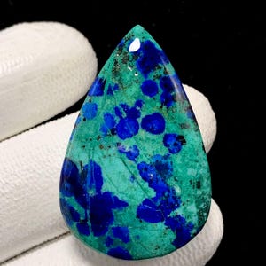 Può includere: Una gemma a forma di goccia, lucidata e dai colori vivaci. Presenta una base turchese e verde acqua, con sorprendenti motivi e macchie blu scuro. La superficie lucida riflette la luce, evidenziando le variazioni naturali e le texture della pietra.