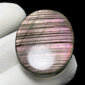 May include: Oval-shaped labradorite gemstone with a polished surface. The stone features a mix of gray, purple, and pink hues with horizontal striations. The gemstone is held against a white background.