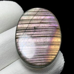 May include: Oval-shaped labradorite gemstone with a polished surface. The stone displays a mix of colors, including purple, pink, and gold, with a striped pattern. The gemstone is held against a white fabric background.