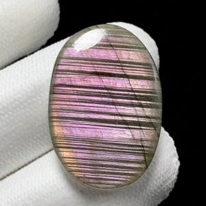 May include: Oval-shaped gemstone with a polished surface. The stone features a pattern of parallel lines in shades of purple, pink, and gray. The gemstone is held against a white background.