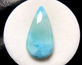 May include: A teardrop-shaped gemstone, showcasing a gradient of light blue to turquoise hues. The polished surface reflects light, highlighting the stone's natural patterns. The gem is set against a white, circular backdrop, with a dark background.