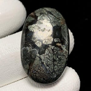 Könnte beinhalten: Ein ovales Schmuckstück mit dunkler, glänzender Oberfläche. Der Stein weist ein Netzwerk aus silbernen und grünen Adern mit Gold- und Silberflecken auf. Der Edelstein wird von einer weißen Oberfläche gehalten.