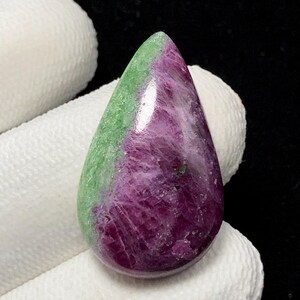 Może przedstawiać: Wypolerowany kamień szlachetny w kształcie łzy. Jedna strona jest w głębokim kolorze magenta, druga w jasnej zieleni. Kamień ma błyszczącą powierzchnię. Kształt kamienia przypomina kroplę wody.