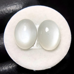 May include: Two oval-shaped, polished moonstone gemstones. The stones are a milky white color with a subtle sheen, set on a white, circular surface. The gemstones are displayed against a dark background.
