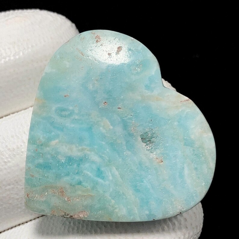 May include: A heart-shaped gemstone in shades of light blue and white. The polished stone displays natural colour variations and textures. The gemstone is held by a white surface against a black background.