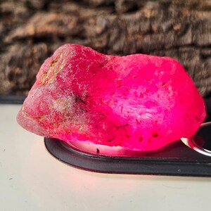 May include: A vivid, raw ruby gemstone in a deep pink shade. The uncut stone has a natural, textured surface with areas of lighter and darker tones. The ruby is set on a black base, highlighting its intense colour.