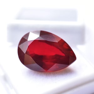 May include: A faceted, pear-shaped, deep red gemstone. The gem is set against a white background, showcasing its brilliant cut and rich colour. The gemstone's facets reflect light, creating a sparkling effect.