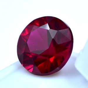 Peut inclure: Une pierre précieuse de type rubis, taillée en forme ovale et à facettes. La gemme est d'un rouge profond, avec de multiples facettes qui réfléchissent la lumière. La pierre est posée sur un fond blanc.