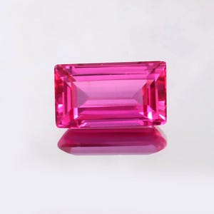 May include: A vibrant, rectangular-cut, hot pink gemstone. The gem is highly reflective, with a polished surface and internal facets that catch the light. The gem is set against a white background, with its reflection visible below.
