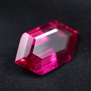 May include: A faceted, rectangular gemstone with a vibrant magenta hue. The gem features a series of precisely cut facets that reflect light, creating a brilliant sparkle. The gemstone is set against a dark background.