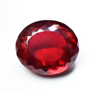 Könnte beinhalten: Ein facettierter, ovalförmiger, tiefroter Edelstein. Der Edelstein hat einen Brillantschliff mit vielen kleinen, dreieckigen Facetten, die das Licht reflektieren. Der Stein ist vor einem weißen Hintergrund platziert, was seine Farbe und Klarheit hervorhebt.
