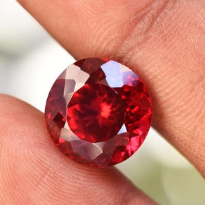 May include: A faceted, round-cut, deep red gemstone. The gem is held between fingers, showcasing its brilliant facets and rich color. The gemstone's polished surface reflects light, creating a sparkling effect. This image highlights the beauty of the gemstone.