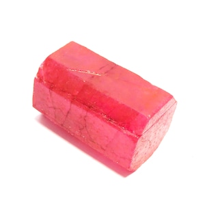 Puede incluir: Una gema de rubí en bruto, facetada y rectangular. El rubí es de un color rojo vibrante y saturado, con texturas e imperfecciones naturales visibles. La gema se encuentra sobre un fondo blanco.