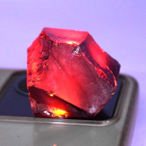 May include: A faceted, deep red gemstone with a rough, natural texture. The gem is illuminated, showcasing its internal structure and color variations. The stone is set against a neutral background.