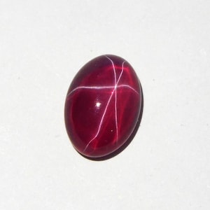 May include: A polished, oval-shaped, deep red gemstone with a star-like pattern of white lines across its surface. The gemstone has a smooth, glossy finish and is set against a white background.