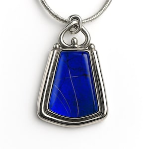 May include: A silver pendant necklace featuring a deep blue, rectangular stone with white veining. The stone is set in a silver frame with a decorative bail and hangs from a silver chain. The pendant has a polished finish.