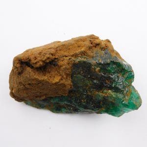 May include: A rough-cut emerald gemstone with a mix of deep green and brown hues. The emerald has a crystalline structure with a rough, uneven surface. The brown portion appears to be a layer of matrix or surrounding rock.