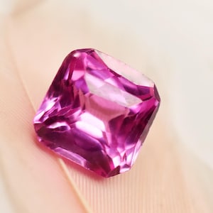Könnte beinhalten: Ein facettierter, quadratisch geschliffener, pinkfarbener Edelstein mit einer brillanten, reflektierenden Oberfläche. Der Edelstein ist vor einem weichen, hellrosa Hintergrund platziert, was seinen Farbton und seine Klarheit hervorhebt. Die Facetten des Edelsteins fangen das Licht ein.
