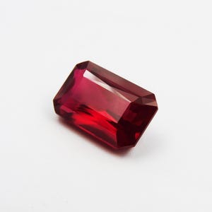 May include: A faceted, rectangular-cut ruby gemstone. The gem is a deep, saturated red color with sharp, clean edges. The gemstone is set against a white background, highlighting its brilliance and clarity.