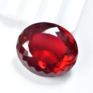 Puede incluir: Una gema facetada, de forma ovalada, de color rojo intenso. La gema tiene un corte brillante con numerosas facetas que reflejan la luz, creando un efecto brillante. La piedra está sobre un fondo blanco.