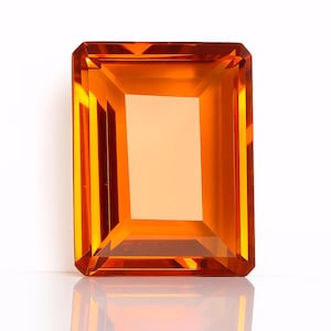 May include: A rectangular, faceted gemstone with a warm orange hue. The gem has a step-cut design, with a large central facet and multiple parallel facets around the edges. The gem is set against a white background, with a slight reflection.