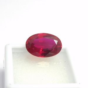 Könnte beinhalten: Ein facettierter, oval geschliffener Rubin-Edelstein. Der Edelstein hat eine tiefe, gesättigte rote Farbe mit einem hohen Reinheitsgrad. Der Rubin ist vor einem weißen Hintergrund platziert, um seine Brillanz und Farbe hervorzuheben. Der Edelstein ist wahrscheinlich für Schmuck.