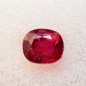 May include: A cushion-cut, faceted ruby gemstone. The gem is a deep, saturated red color with multiple facets that reflect light. The gemstone is set against a white background.
